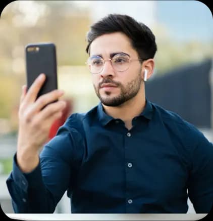 A man with a beard and glasses is taking a selfie with a smartphone. He is wearing a dark blue shirt and wireless earphones.